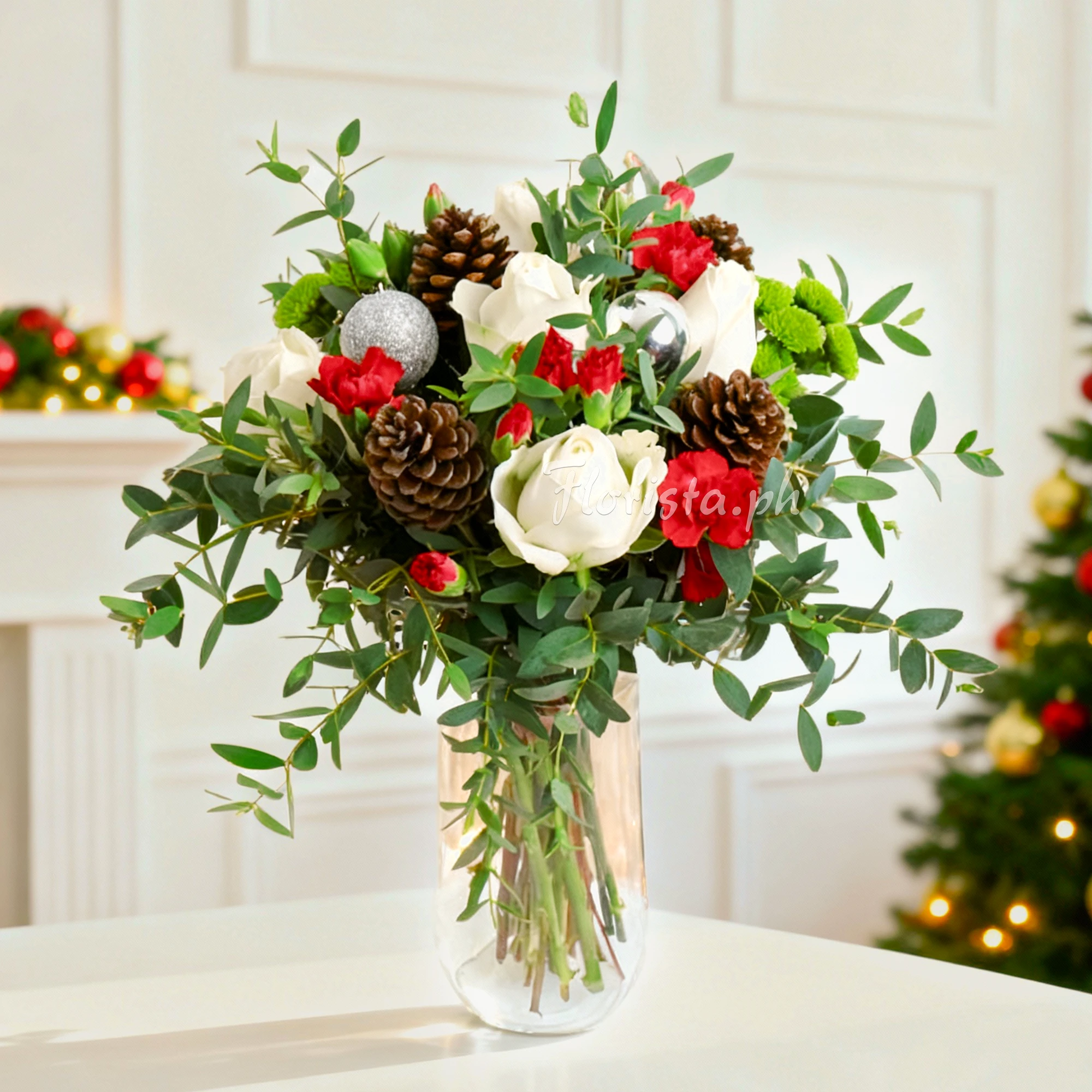 An arrangement of 5 white roses, 3 red spray carnations, 4 pinecone, 3 green button chrysanthemums, silver ornaments with green fillers in a glass vase.